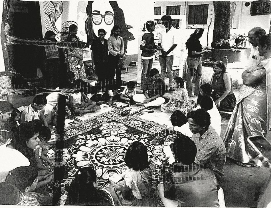 An art-making circle: adults and children seated on a mat on the ground with paper, paint and pencils, under a tree.